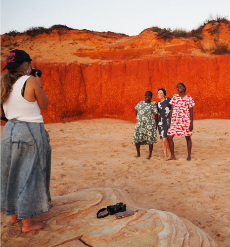 A photographer stands barefoot on sandy ground, taking a photo of three women standing together in front of a striking red cliff face. The women are smiling and wearing patterned dresses, framed by the warm tones of the landscape, suggesting a relaxed, collaborative moment outdoors.